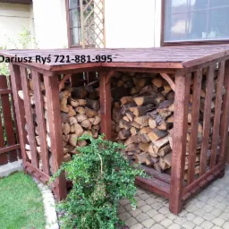 Drewniana wiata na drewno kominkowe, wypełniona pociętymi kawałkami drewna, z ciemnobrązowym dachem i ściankami, ustawiona na kostce brukowej obok trawnika i ogrodzenia.