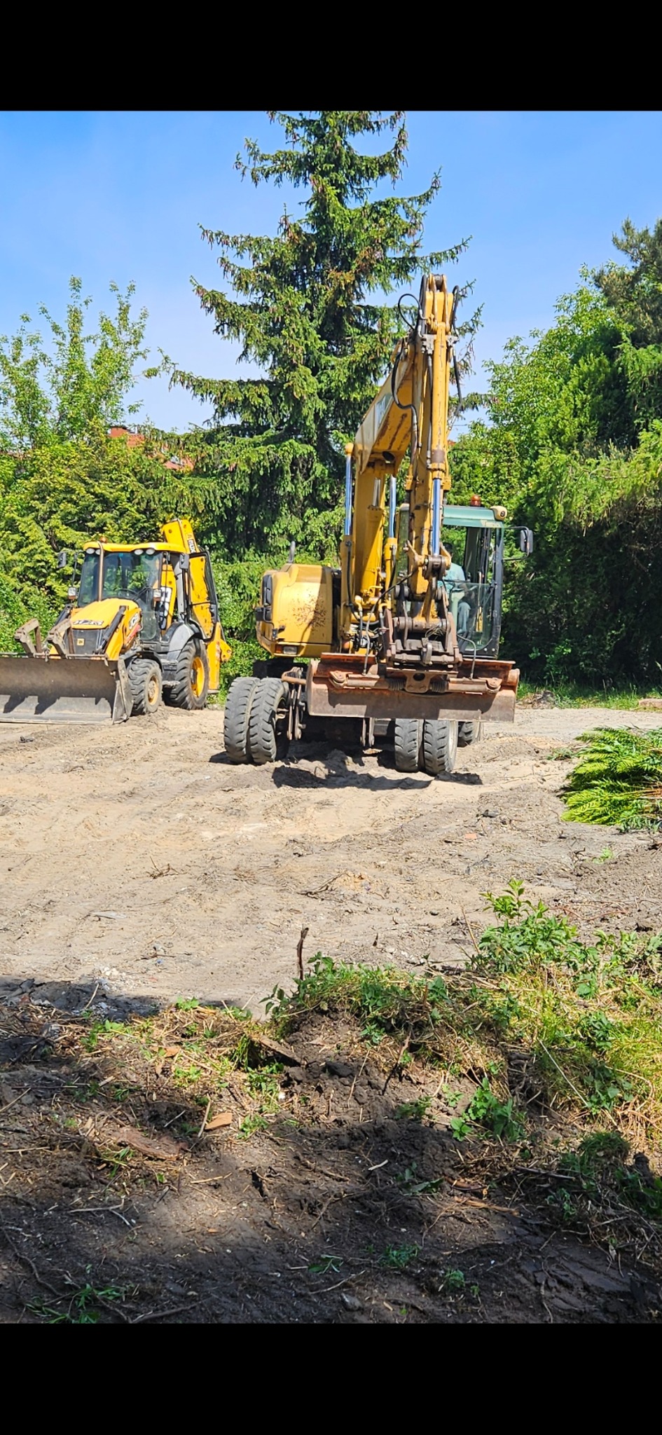 Żółta koparka kołowa i koparko-ładowarka na terenie budowy, widoczne ślady prac ziemnych i wycinki, w tle drzewa iglaste i liściaste pod błękitnym niebem.