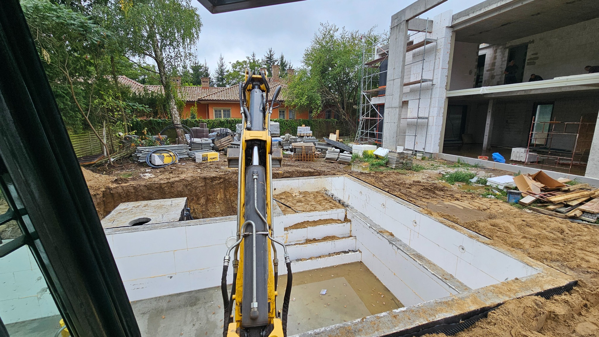 Żółta koparka z widokiem na wykopany basen z betonowymi ścianami i schodami, w tle budynek w trakcie budowy i domy jednorodzinne.