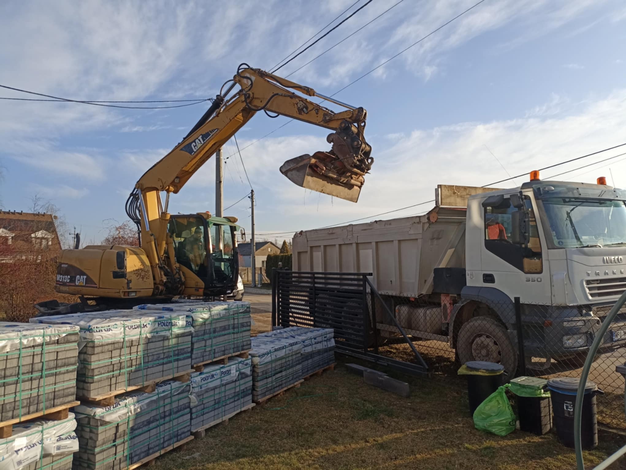 Żółta koparka CAT nabiera ziemię do wywrotki IVECO, obok palety z kostką Polbruk na trawie, w tle domy i błękitne niebo.