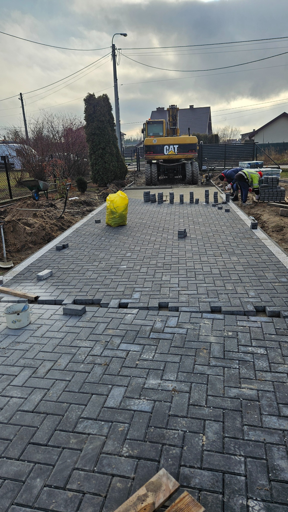 Montaż szarej kostki brukowej w jodełkę na posesji, z widoczną koparką CAT w tle i pracownikami układającymi obrzeża.