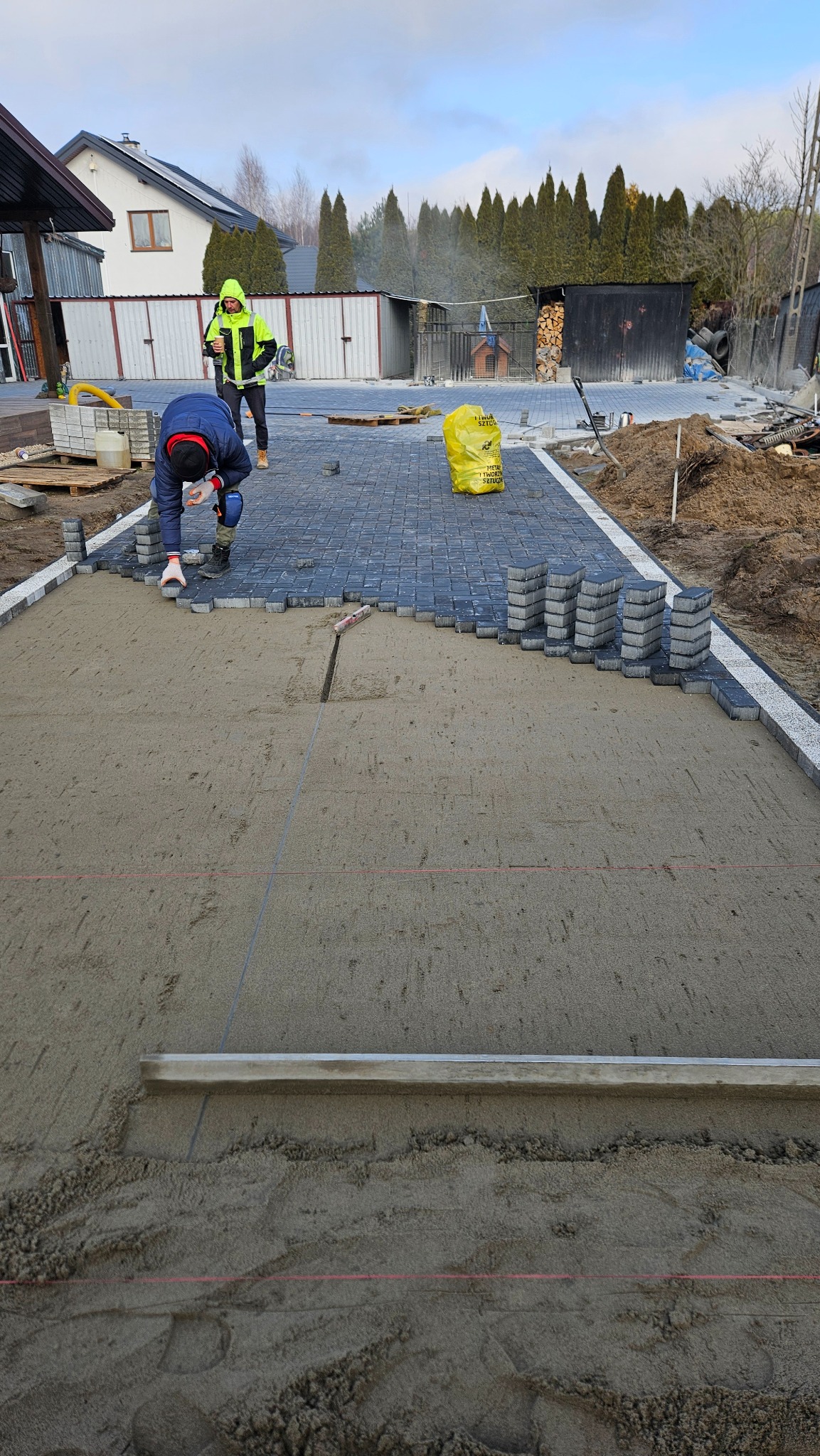 Układanie szarej kostki brukowej na podsypce piaskowej, widoczni dwaj pracownicy, jeden w odblaskowej kurtce, w tle budynki gospodarcze i drzewa iglaste.