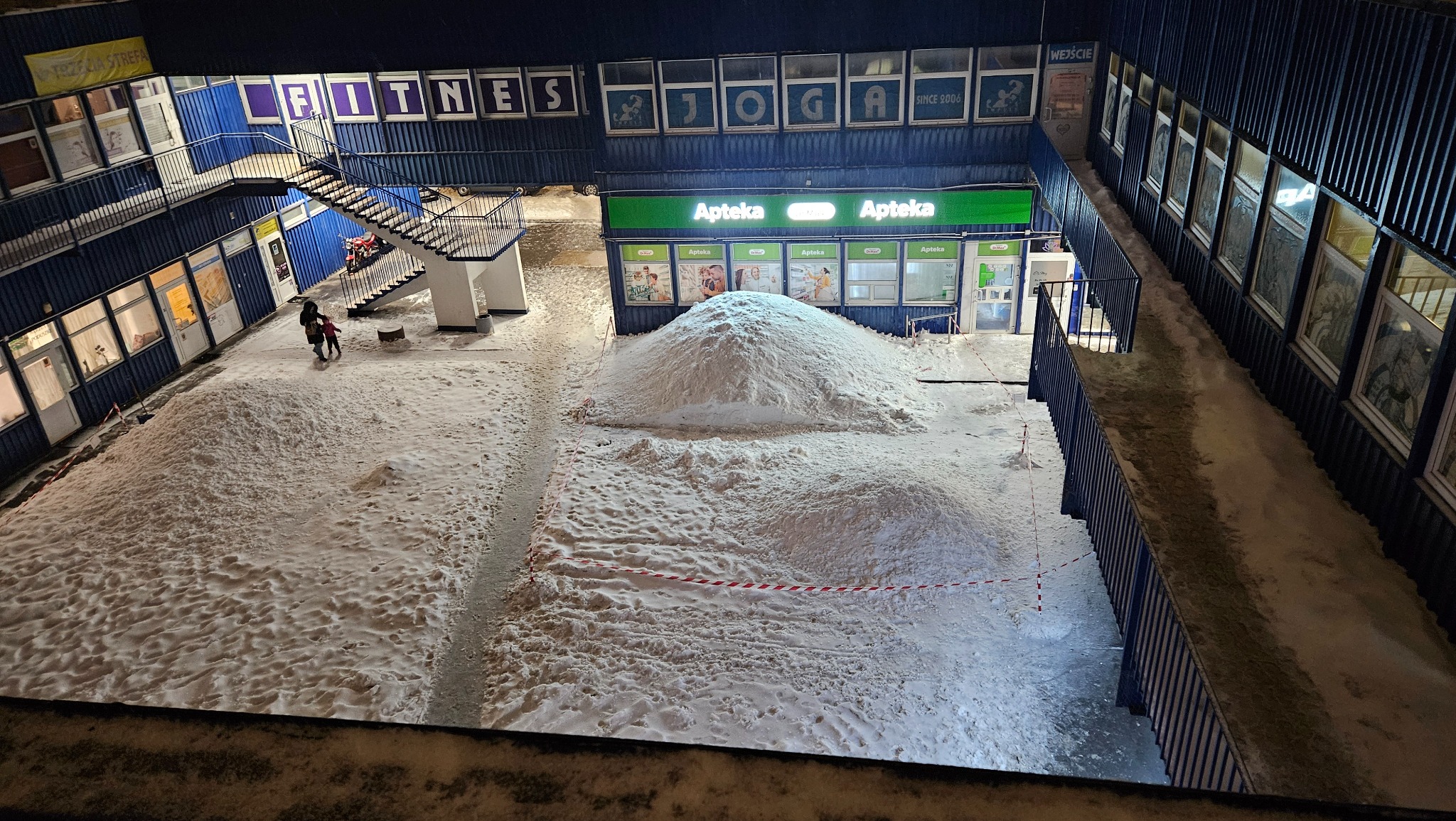 Zasypane śniegiem wejście do apteki w budynku z napisem FITNESS i JOGA. Śnieg tworzy duże pryzmy, teren odgrodzony taśmą. Widoczna osoba z dzieckiem.
