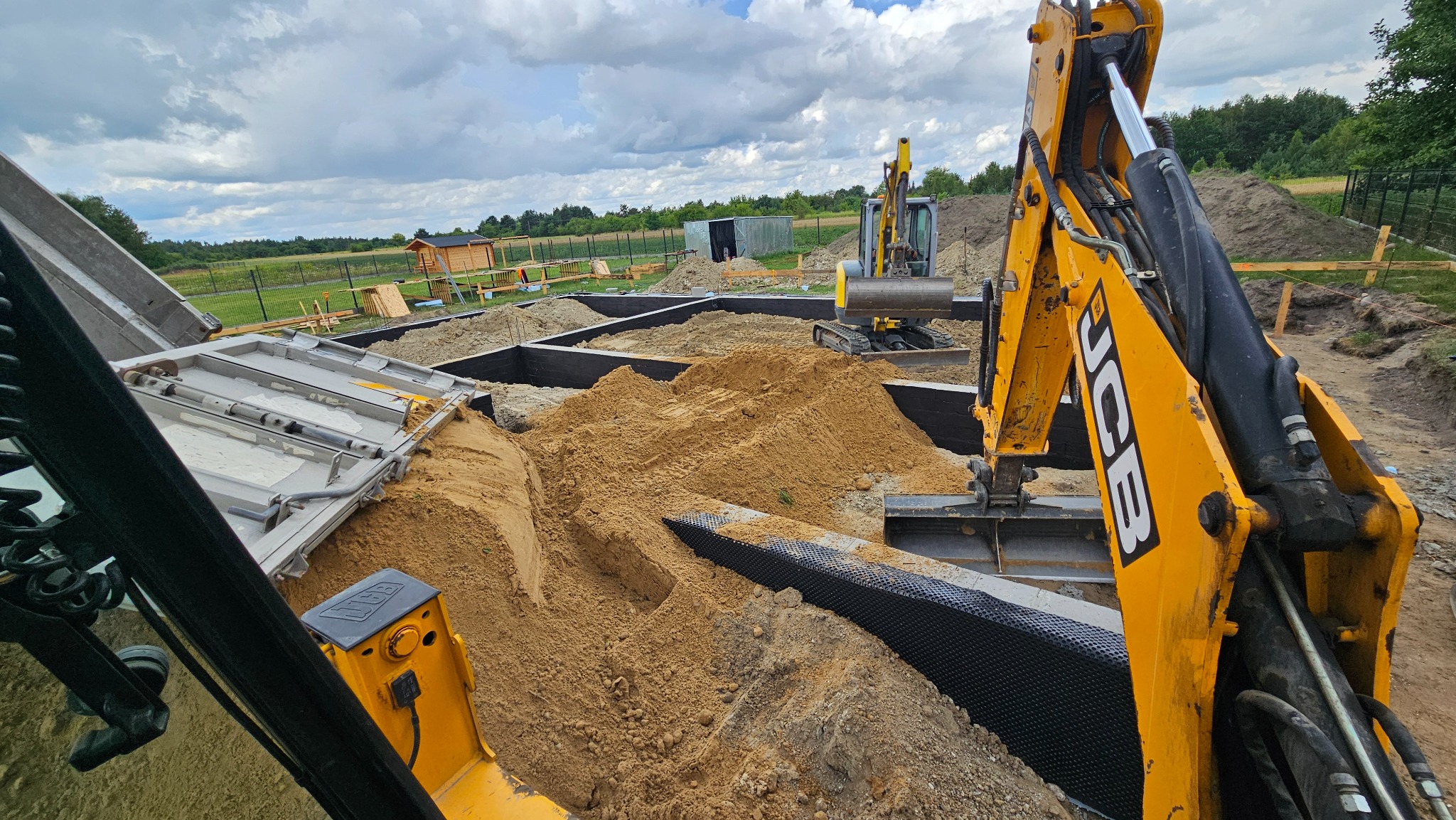 Żółta koparka JCB zasypuje piaskiem wykop pod fundamenty z izolacją, w tle widoczna inna koparka i częściowo zasypane fundamenty. Pochmurne niebo.