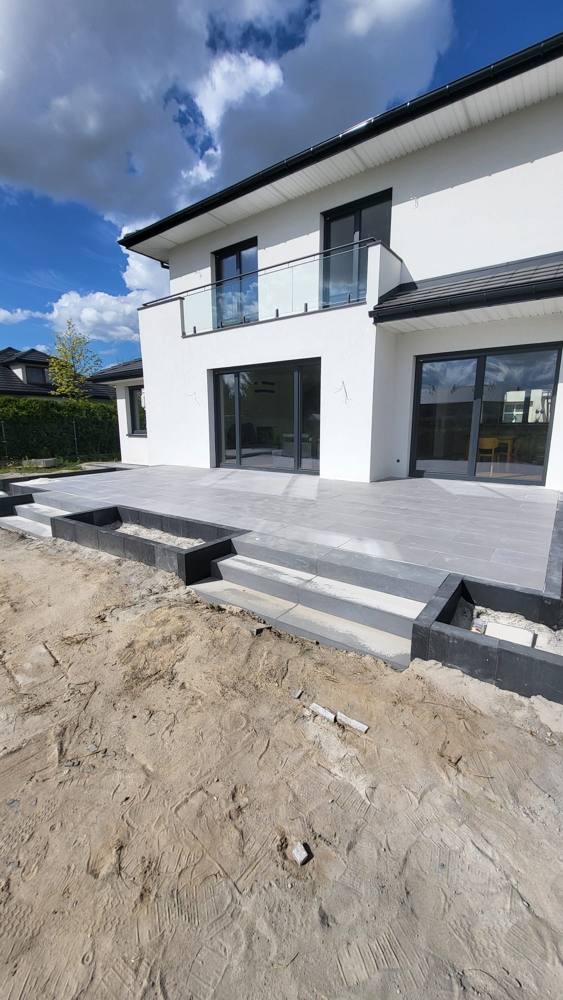 Nowoczesny taras z szarych płyt, z niskimi schodami i obrzeżami z ciemnych kostek. W tle biały dom z dużymi oknami i balkonem. Częściowo wykończony teren wokół tarasu.