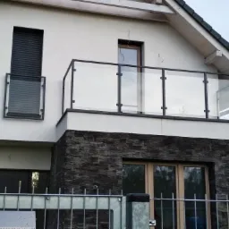 Nowoczesny balkon z czarną balustradą szklaną, widoczny zza metalowego ogrodzenia, z elewacją budynku wykończoną kamieniem łupkowym.