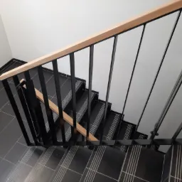 Klatka schodowa z czarnymi płytkami i drewnianą poręczą, widok z góry, minimalistyczny design.