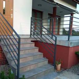 Zewnętrzne schody wejściowe z szarymi płytkami i nowoczesną metalową balustradą z poziomych prętów, prowadzące do drzwi wejściowych domu z czerwonymi ścianami i widocznym fragmentem tarasu.