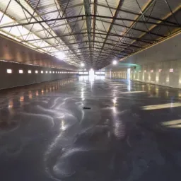 Błyszcząca, ciemna posadzka w przestronnym, betonowym wnętrzu hali z metalową konstrukcją dachu i rzędem okien w górnej części ścian. Widoczne refleksy światła.