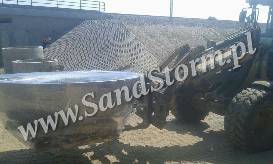 Ciężka maszyna transportuje okrągły, zabezpieczony element na tle betonowej konstrukcji i ażurowych płyt. Widoczny operator i betonowe kręgi.