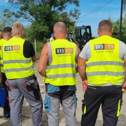 SzkoleniaMM Rafał Dunaj - Grupa mężczyzn w kamizelkach odblaskowych z logo 'Szkolenia MM' obserwuje pracę koparki na placu budowy. Błękitne niebo i drzewa w tle. Widok z tyłu.