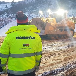SzkoleniaMM Rafał Dunaj - Instruktor w żółtej kurtce Komatsu na tle pracującego spychacza i grupy obserwatorów w zimowym krajobrazie. Szkolenie z obsługi maszyn budowlanych.