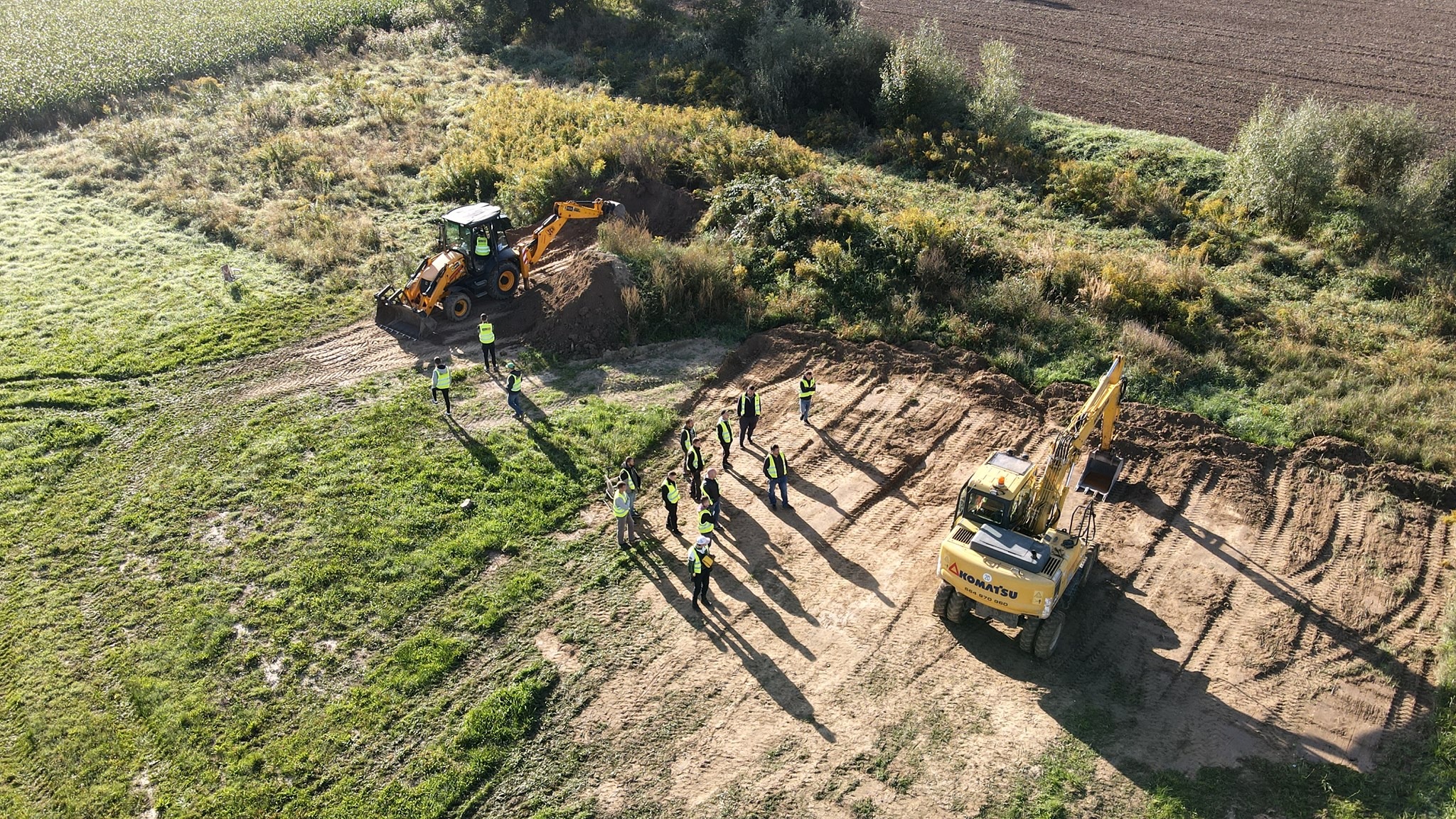 Zajęcia praktyczne z obsługi koparki jednonaczyniowej i koparkoładowarki, widok z góry na grupę kursantów w kamizelkach odblaskowych i dwie maszyny budowlane na placu manewrowym.