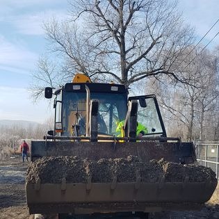 Żółta koparkoładowarka z otwartymi drzwiami kabiny i łyżką wypełnioną ziemią, na placu budowy w słoneczny dzień. W tle widoczny pracownik w czerwonej kurtce i drzewa bez liści.