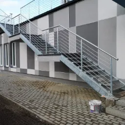 Zewnętrzne, metalowe schody z balustradą prowadzące na pierwsze piętro budynku o szaro-białej elewacji. Na dole widoczne wiadro po farbie i świeżo ułożona kostka brukowa.