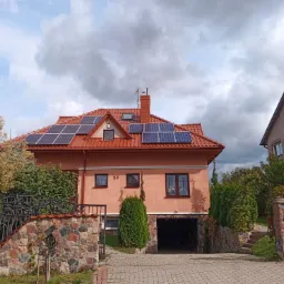 Dom jednorodzinny z czerwoną dachówką i panelami słonecznymi na dachu, widoczny z poziomu ulicy, z bramą wjazdową i bujną roślinnością w tle.