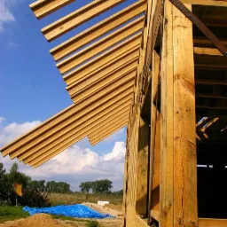 Drewniany szkielet domu w budowie, widok z dołu na konstrukcję dachu i ściany, z niebieską plandeką w tle. Widoczne elementy złączne i naturalne drewno.