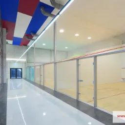 Nowoczesne wnętrze kortu do squasha z przeszklonymi ścianami, kolorowym sufitem i błyszczącą podłogą. Widoczne detale wykończenia i oświetlenie.