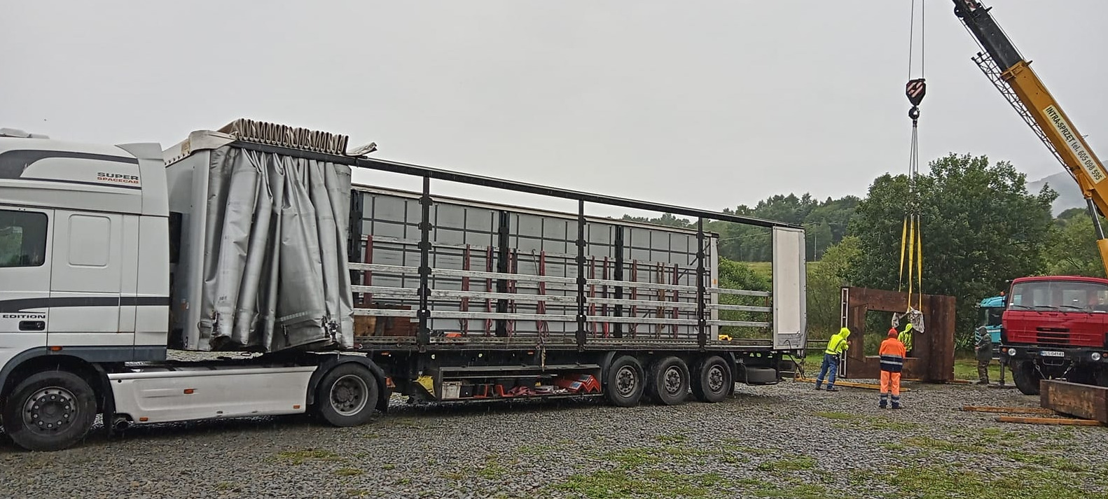 Ciężarówka z otwartą naczepą przewożąca elementy konstrukcyjne, pracownicy w odblaskowych ubraniach przy użyciu dźwigu rozładowują ładunek na żwirowym placu.