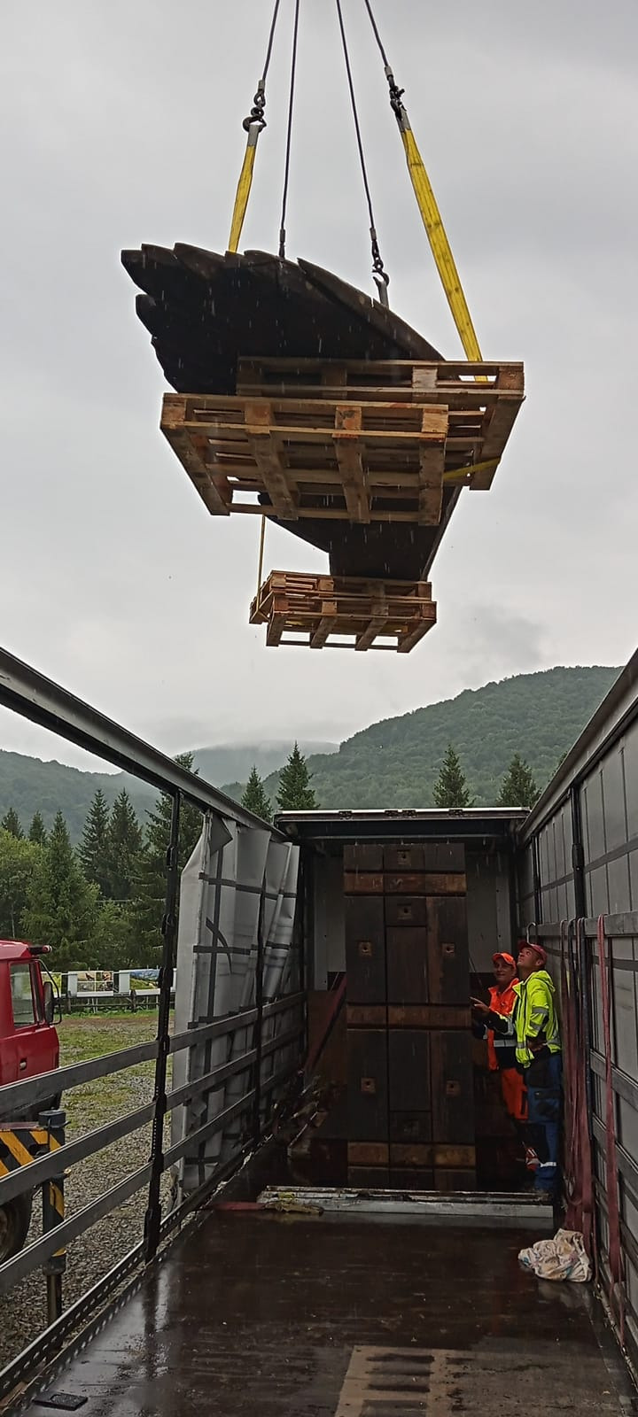 Załadunek elementów drewnianych, przypominających fragmenty statku, na paletach do ciężarówki, przy użyciu pasów transportowych, w tle górzysty krajobraz i pracownicy w odblaskowych ubraniach.
