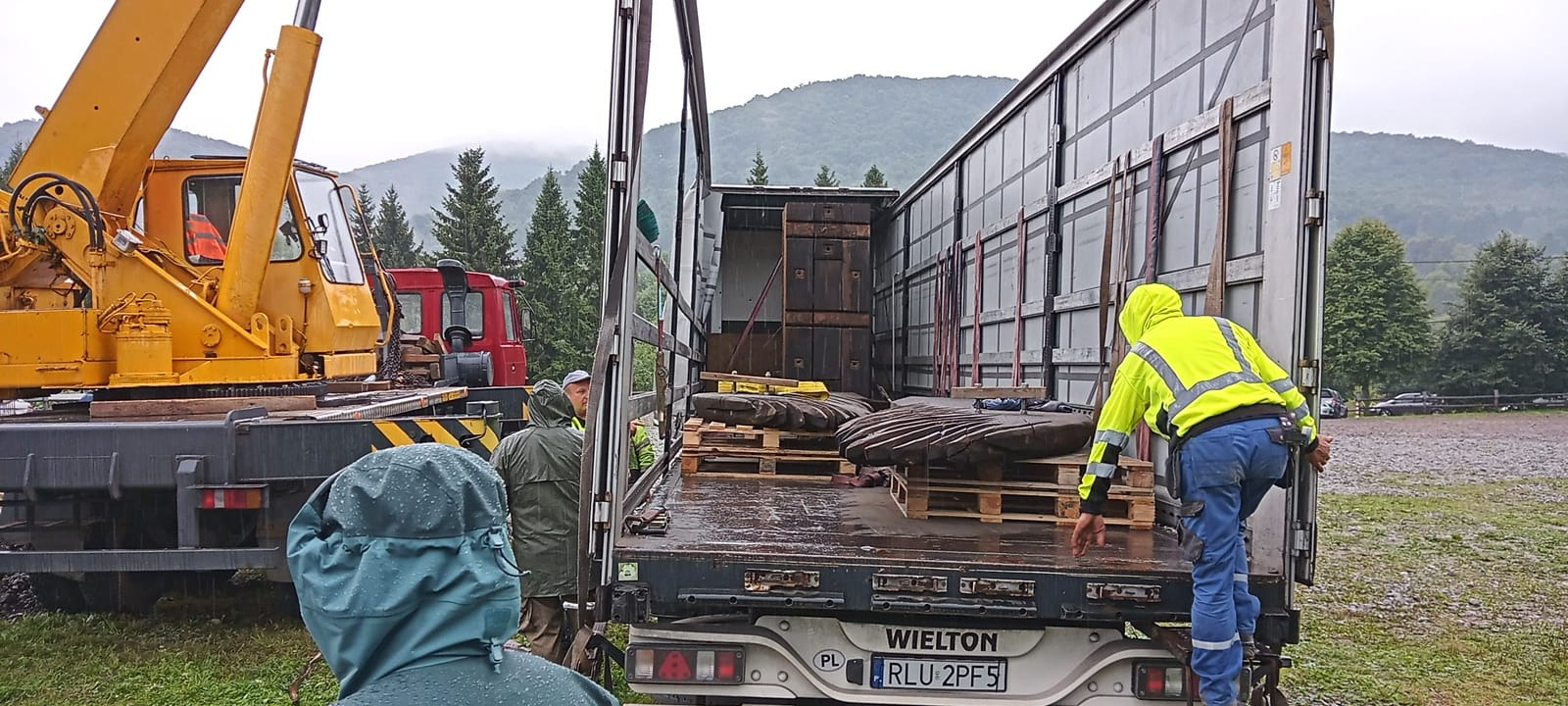 Załadunek ciężkich, okrągłych elementów z wyraźnymi śladami obróbki na paletach do naczepy ciężarówki przy użyciu żółtego dźwigu, widoczni pracownicy w odblaskowych ubraniach i pelerynach...