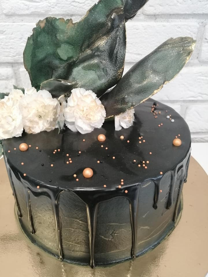 Tort z czarną polewą, zdobiony białymi kwiatami, złotymi kropkami i elementami imitującymi liście w odcieniach zieleni i złota, na tle białej cegły.