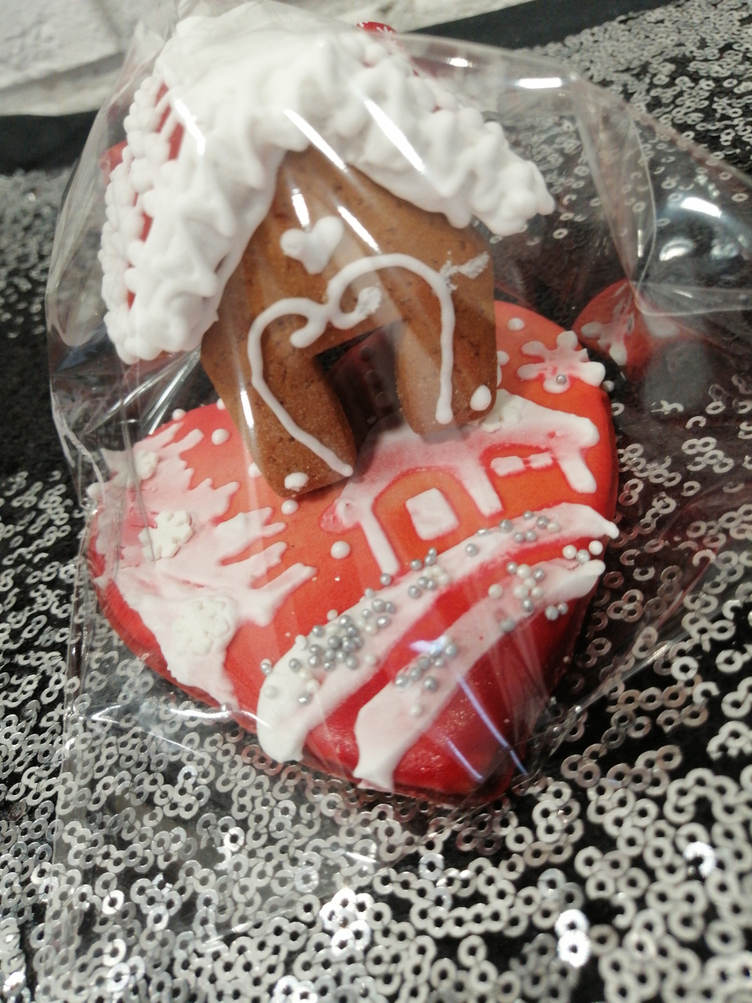 Piernikowy domek z lukrowanymi detalami, umieszczony na piernikowym sercu, zapakowany w przezroczysty celofan, na błyszczącym, srebrnym tle.