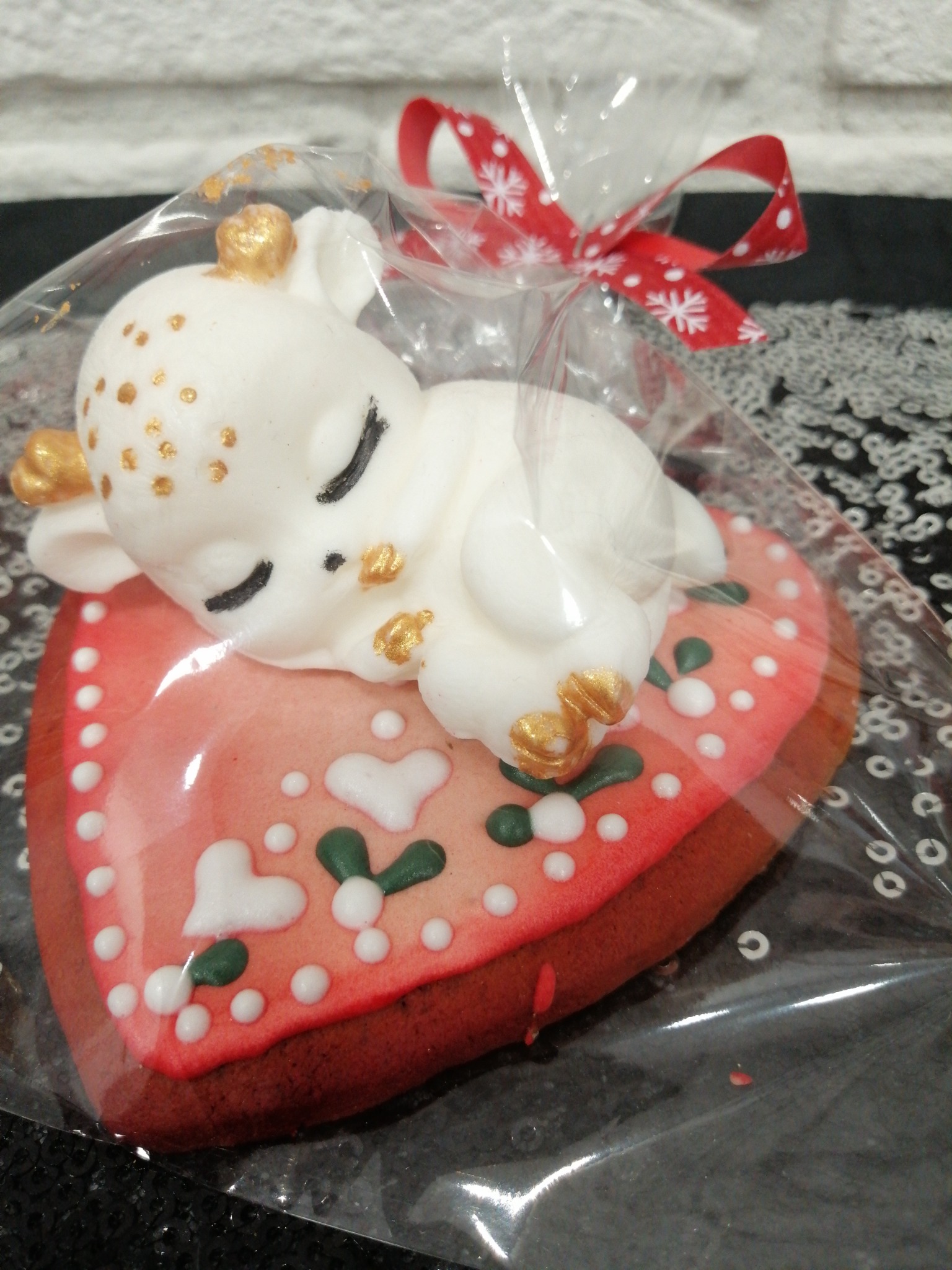 Piernikowe serce z dekoracją przedstawiającą śpiącego jelonka z lukru, zapakowane w przezroczystą folię z czerwoną kokardą w śnieżynki.