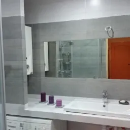 Nowoczesna łazienka w szarościach z kabiną prysznicową, umywalką i lustrem, akcentowana fioletowymi świecami. Widoczny fragment pralki.