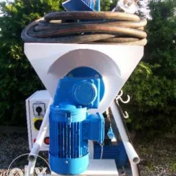 Biało-niebieska maszyna do natrysku tynku z wężem na tle zieleni, widoczny silnik i panel sterowania, umieszczona na zewnątrz.