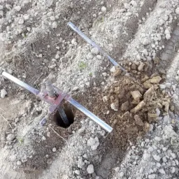 Wiertnica ręczna zagłębiona w ziemi, obok wykopana ziemia i pręty. Widoczne ślady po oponach na polu.