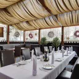 Uroczysta aranżacja wnętrza restauracji z długim stołem, krzesłami i dekoracyjnym sufitem z materiału. Widoczne dekoracje okienne i subtelne oświetlenie.