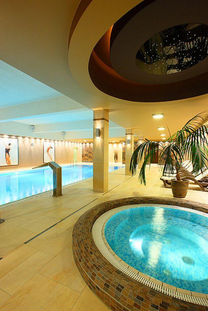 Ekskluzywny basen kryty z jacuzzi, oświetlony nastrojowo, z palmą w donicy i dekoracyjnym sufitem. W tle obrazy na ścianach i leżaki.
