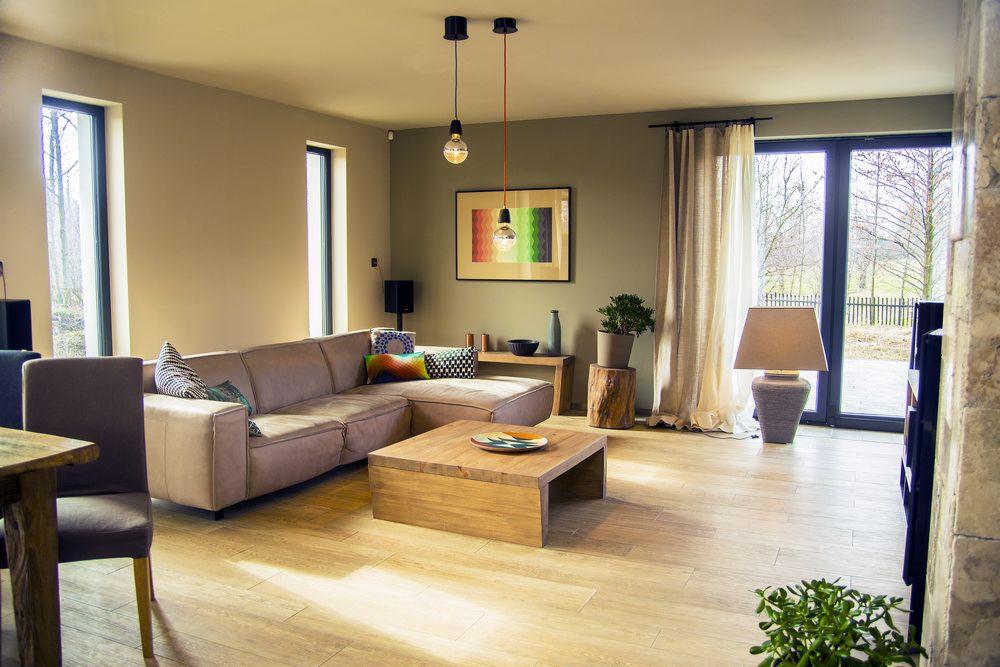 Jasny salon z beżową sofą narożną, drewnianym stolikiem kawowym i parkietem. Dwa okna, lampy wiszące i grafika na ścianie dopełniają wystrój. Widok na zieleń za oknem.