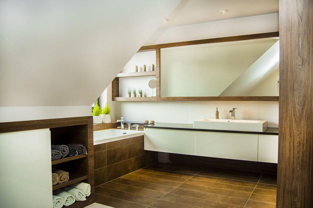 Nowoczesna łazienka na poddaszu z brązowymi płytkami, białą umywalką i dużym lustrem w drewnianej ramie. Minimalistyczne półki dekoracyjne z roślinami.