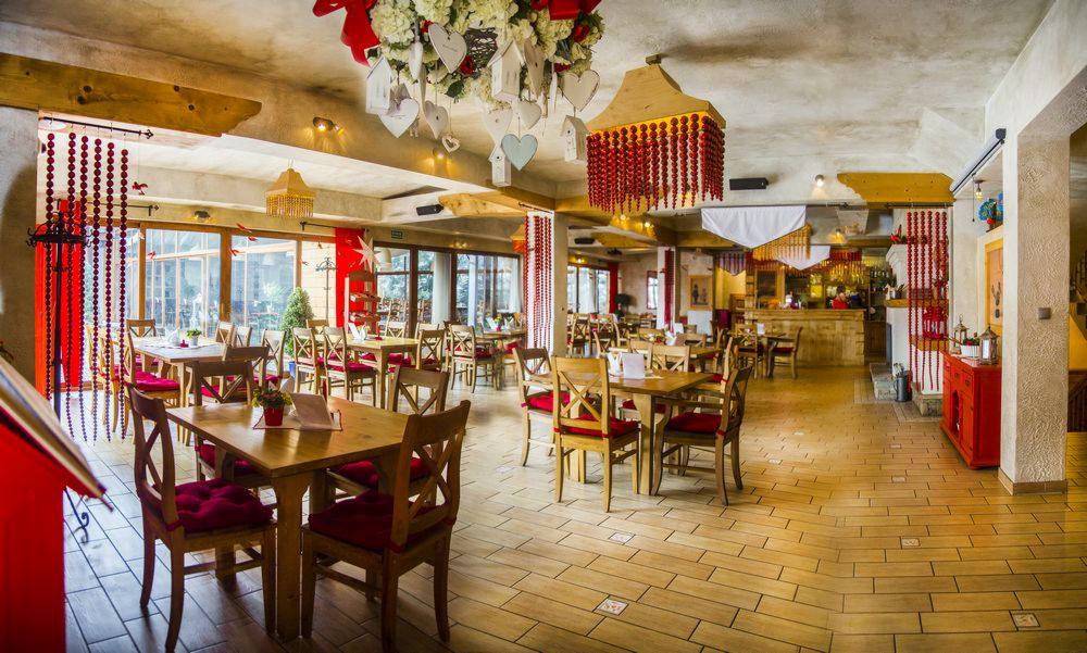Rustykalne wnętrze restauracji z drewnianymi stołami i krzesłami z czerwonymi poduszkami. Dekoracje z kwiatów i serc podwieszone pod sufitem. Ciepła kolorystyka.