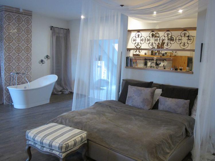 Eklektyczna sypialnia z wanną wolnostojącą, oddzielona zwiewną zasłoną. Nad łóżkiem dekoracyjne półki. Na podłodze szara podłoga imitująca drewno.