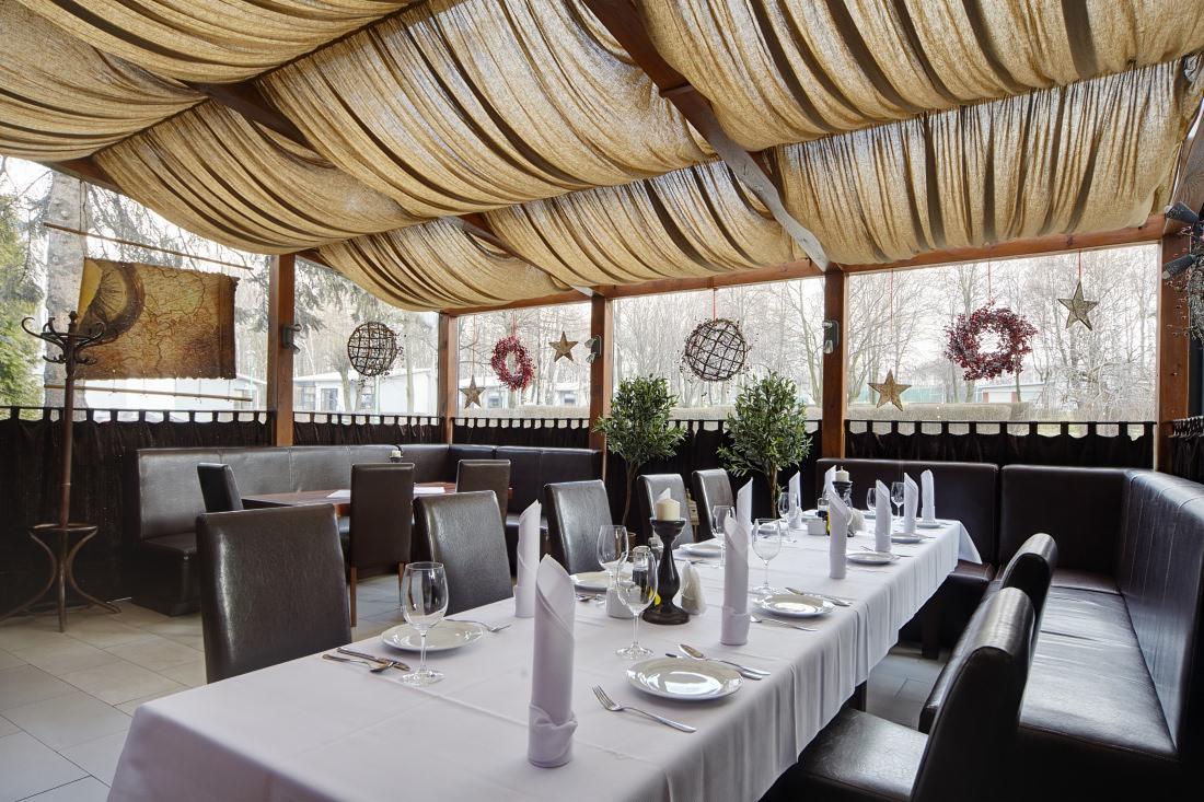 Uroczysta aranżacja wnętrza restauracji z długim stołem, krzesłami i dekoracyjnym sufitem z materiału. Widoczne dekoracje okienne i subtelne oświetlenie.