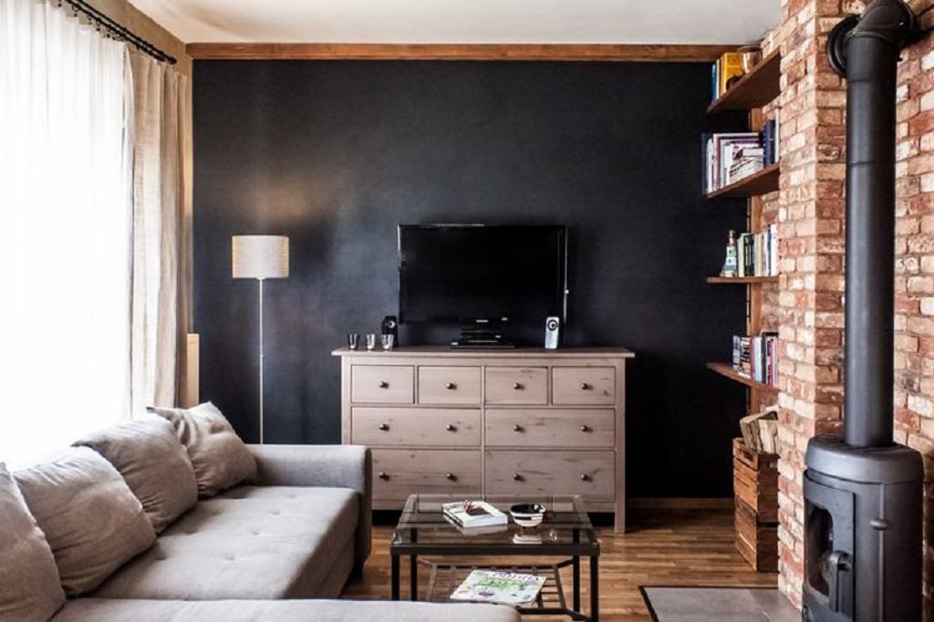 Salon w stylu loft z ceglaną ścianą, kominkiem i drewnianymi półkami. Ciemna ściana za telewizorem, jasna komoda i szara kanapa tworzą przytulne wnętrze.