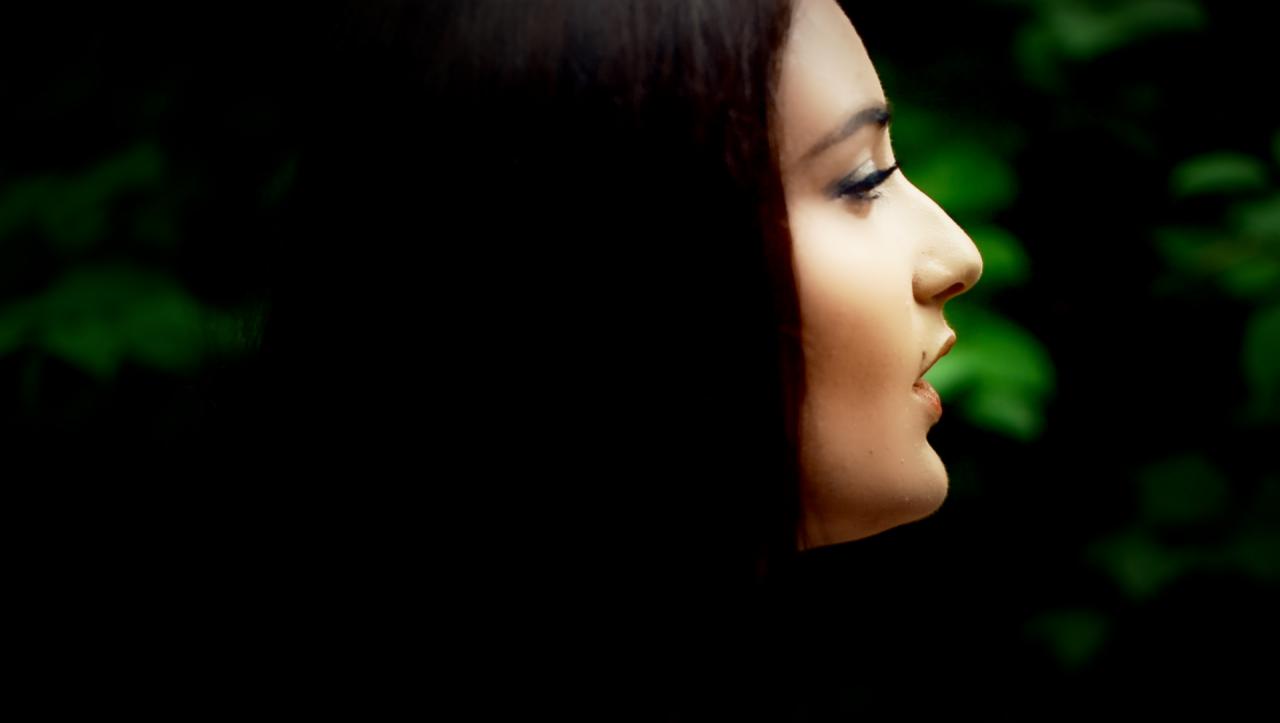 Portret kobiety z profilu, z delikatnym makijażem oczu i ust, na tle ciemnej zieleni liści.