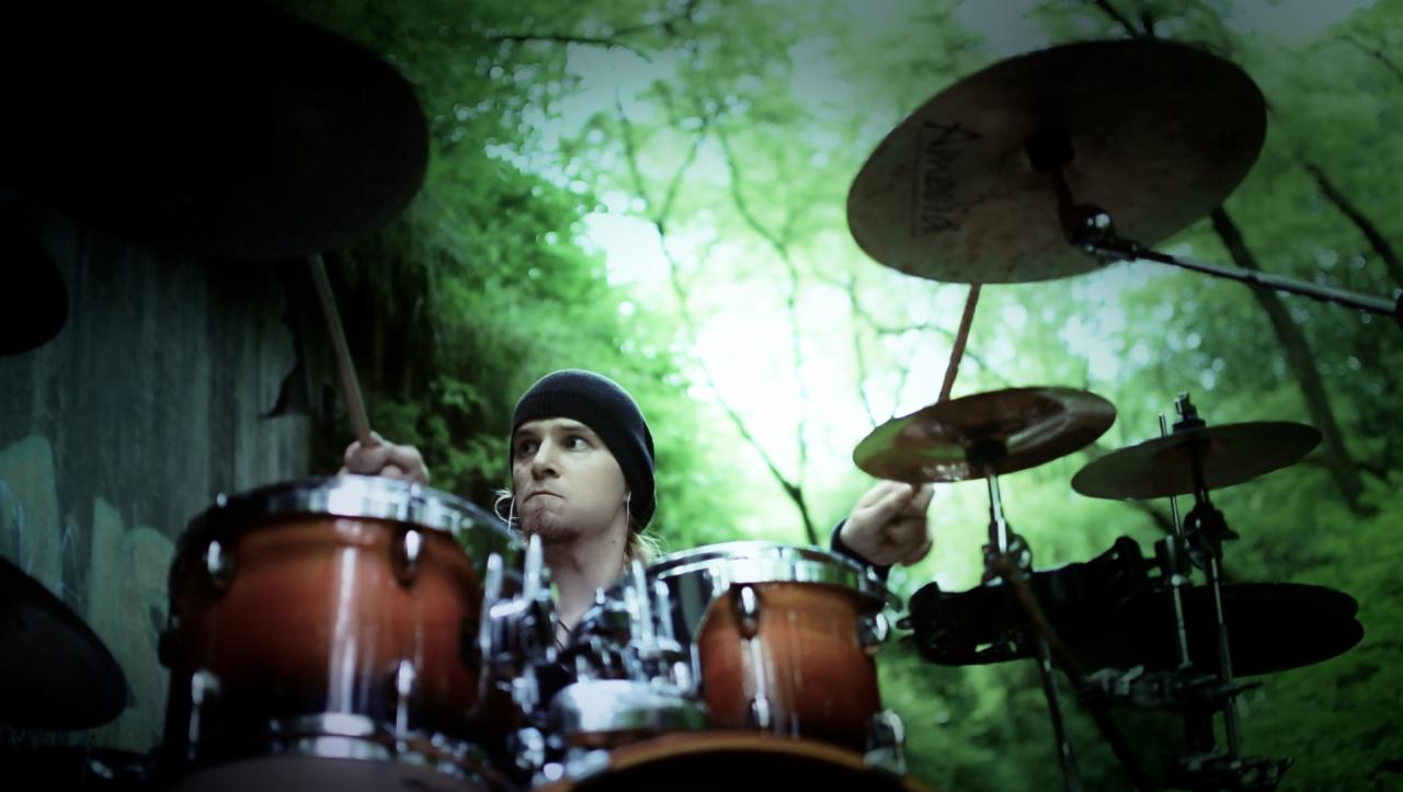 Młody perkusista w czapce gra na perkusji na tle zielonego lasu, ujęcie z niskiej perspektywy.
