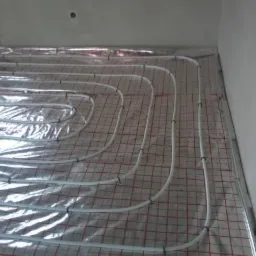 Białe rury ogrzewania podłogowego, ułożone na folii termoizolacyjnej z czerwoną siatką, równomiernie rozłożone w pomieszczeniu przed wylaniem posadzki.