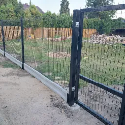 Nowoczesne ogrodzenie z paneli z bramą w kolorze antracytowym na betonowej podmurówce, widok z zewnątrz, w tle drewniane ogrodzenie sztachetowe i zielona roślinność.