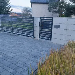Nowoczesna, przesuwna brama metalowa z poziomymi szczeblami, zamontowana na betonowych słupkach, z widocznym automatem do bramy i kostką brukową na podjeździe, otoczona trawą ozdobną.