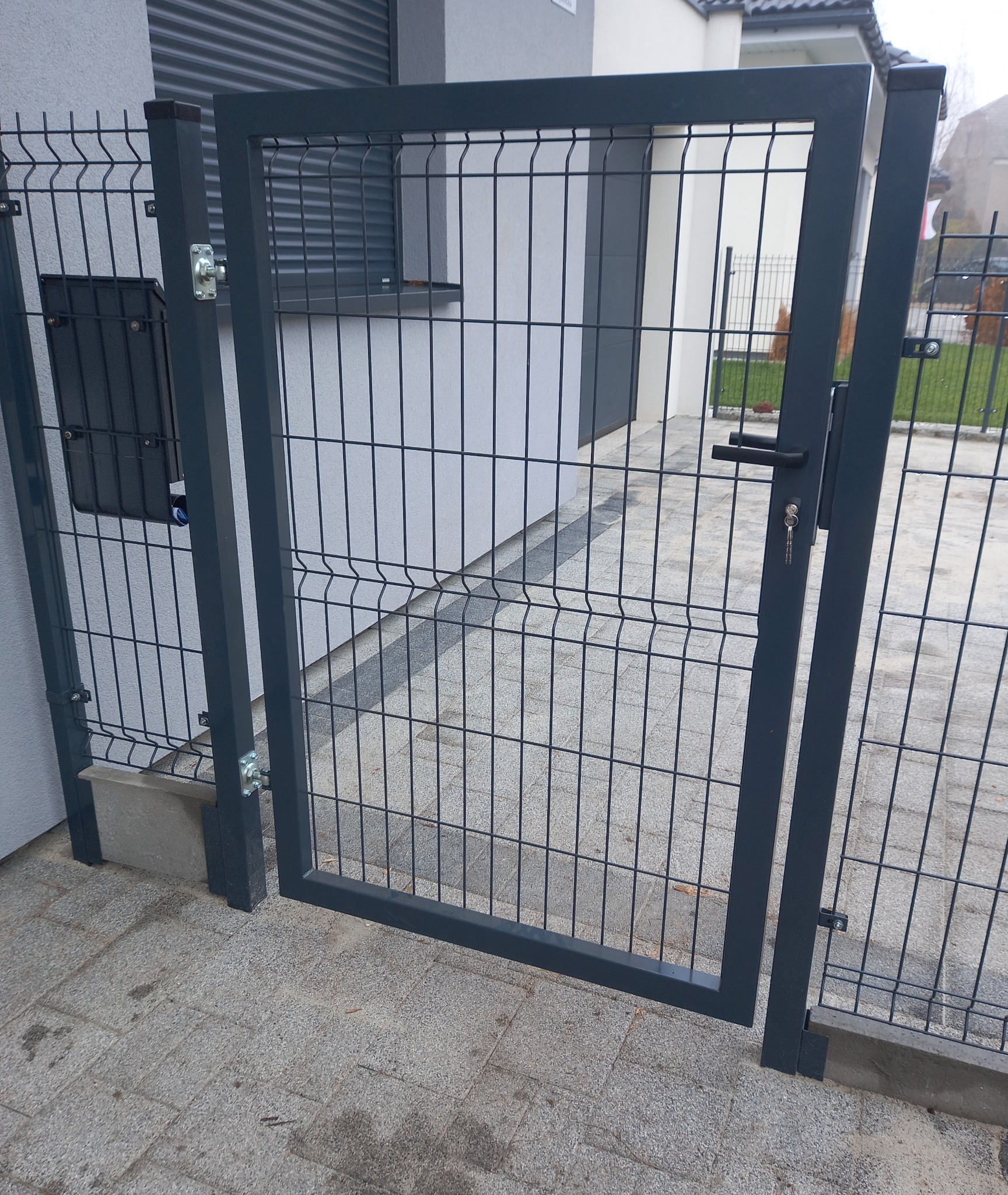 Nowoczesna, grafitowa furtka z paneli z kluczem w zamku, wkomponowana w ogrodzenie panelowe na tle nowego domu.