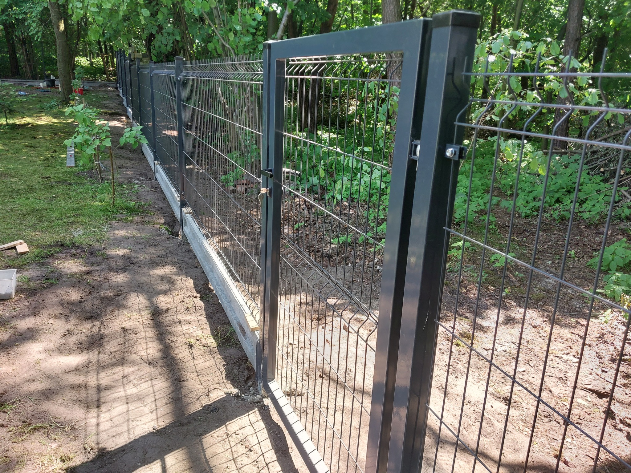 Nowoczesne, antracytowe ogrodzenie panelowe z bramką, osadzone na betonowej podmurówce, w otoczeniu zieleni.