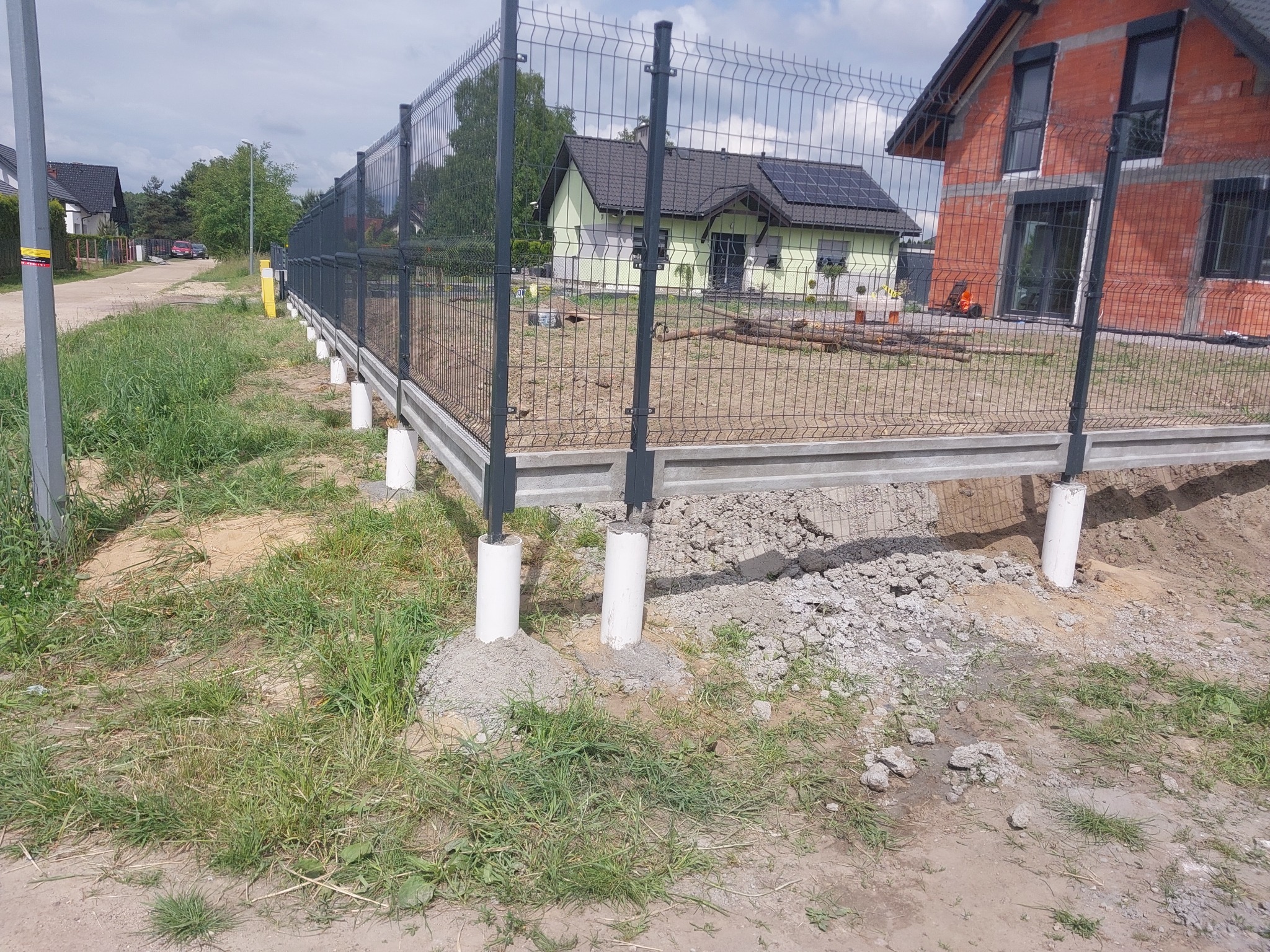 Montaż ogrodzenia panelowego: widok na fragment nowo postawionego ogrodzenia z siatki panelowej na betonowej podmurówce z okrągłymi słupkami, w tle dom w trakcie budowy i dom z panelami słonecznymi.