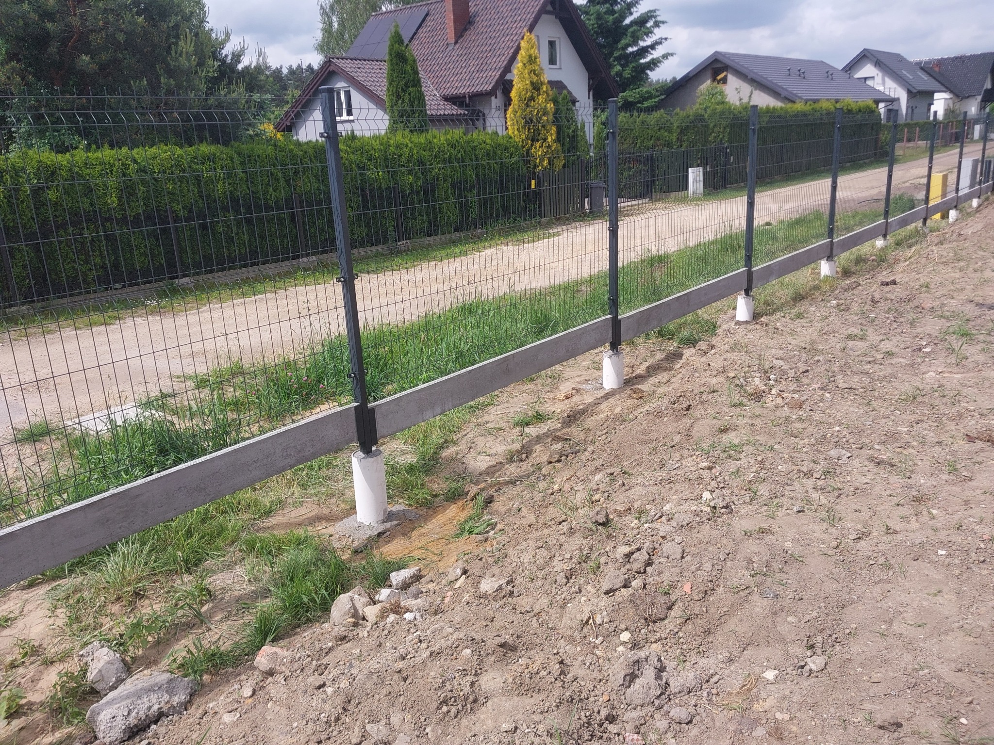 Wznoszenie ogrodzenia z siatki na betonowej podmurówce, widoczne słupki i fragment siatki, w tle domy jednorodzinne.