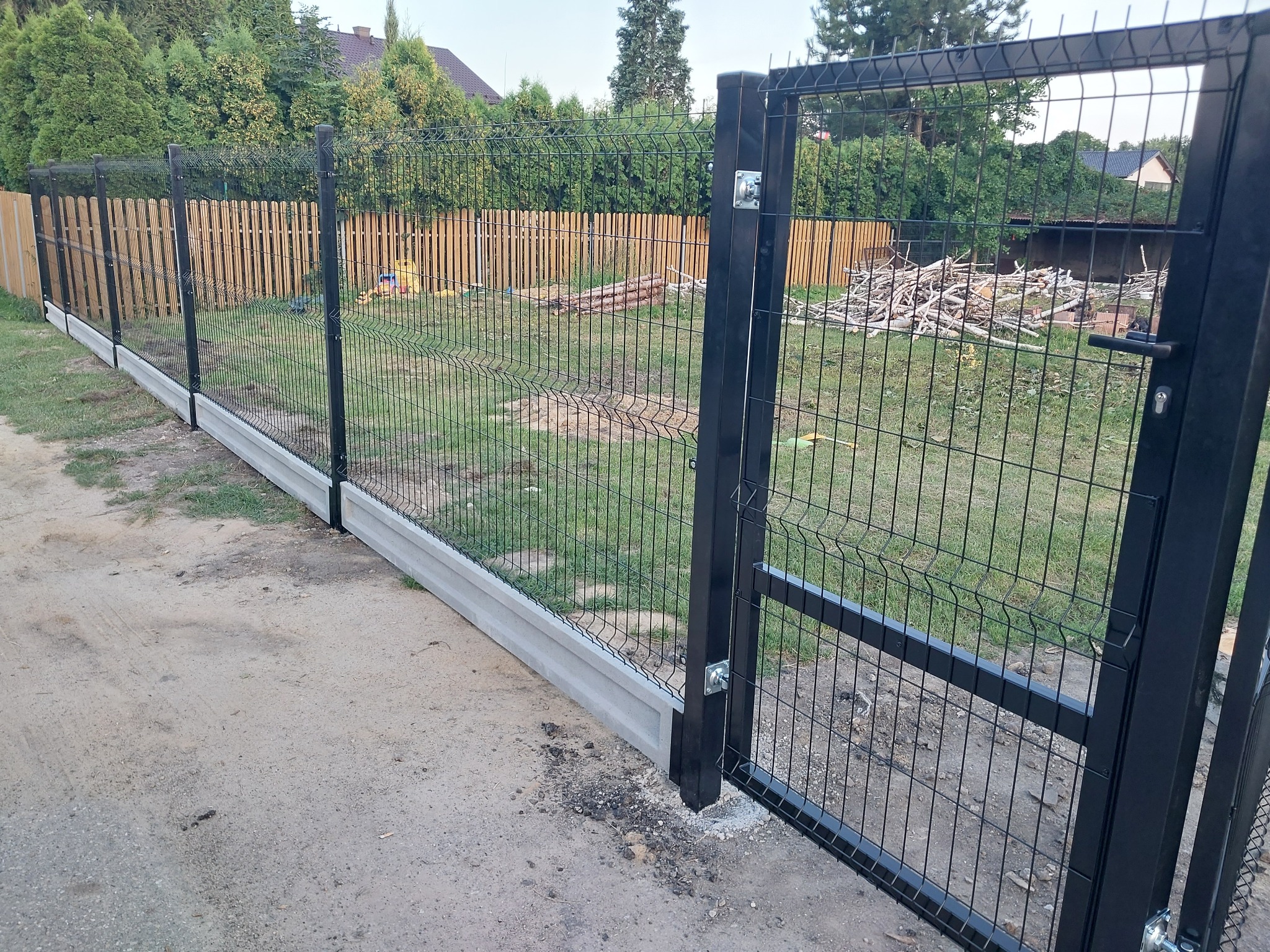 Nowoczesne ogrodzenie z paneli z bramą w kolorze antracytowym na betonowej podmurówce, widok z zewnątrz, w tle drewniane ogrodzenie sztachetowe i zielona roślinność.