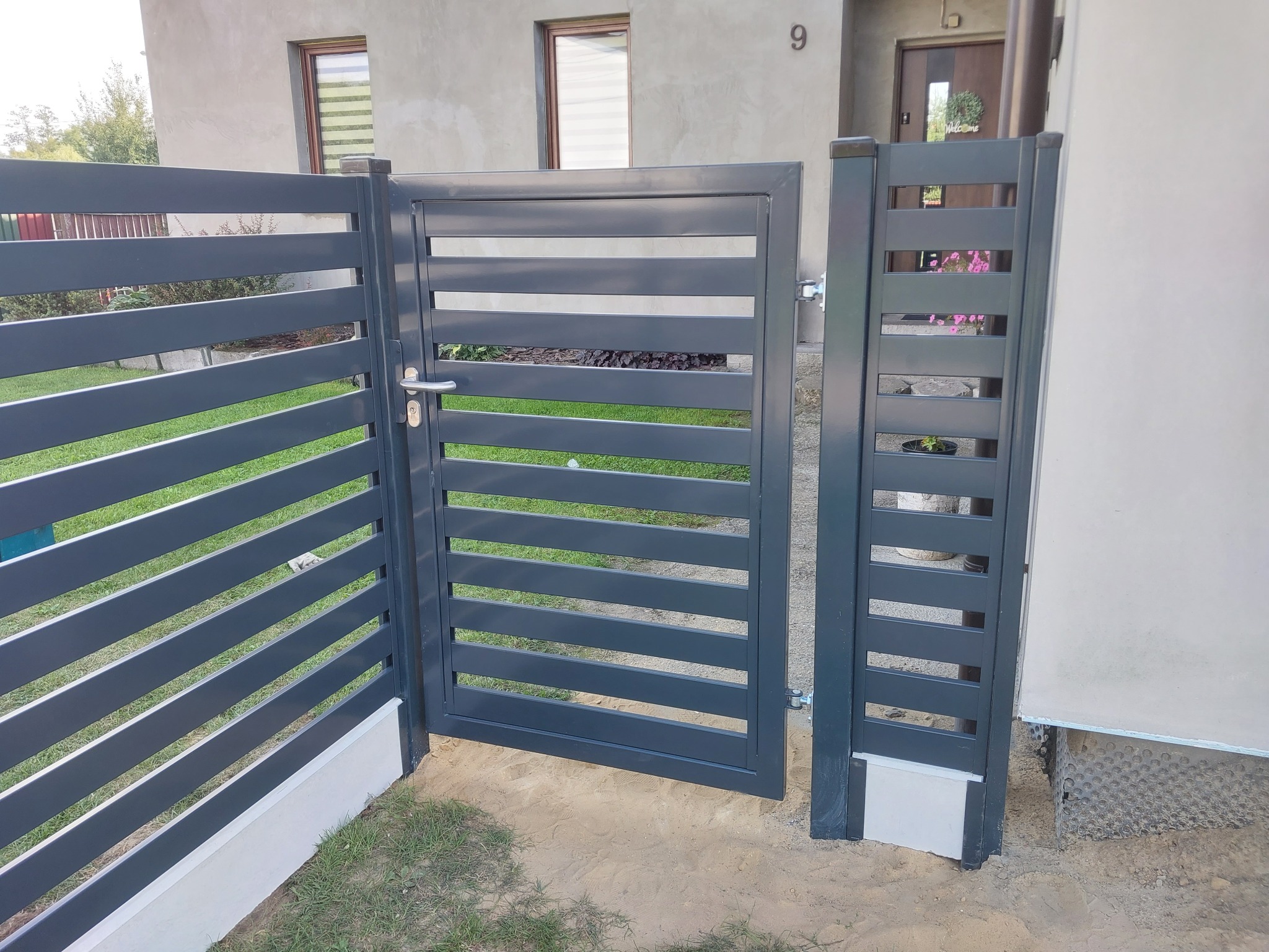 Nowoczesna, grafitowa brama posesyjna z poziomymi panelami, zamontowana przy domu z numerem 9, widok z zewnątrz.